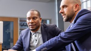 Two men in blazers are sitting closely and looking intently at a computer screen in a bright room.