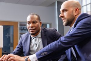 Two men in blazers are sitting closely and looking intently at a computer screen in a bright room.