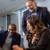 Three people in business attire smile and collaborate while looking at a laptop in a bright office.