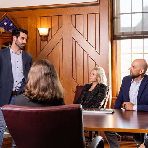 A man in a blazer stands and gestures while three seated people listen attentively in a wood-paneled room.