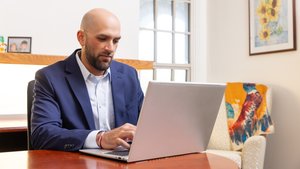 A man in a blue blazer types on a laptop at a wooden table in a room with a window and framed sunflower painting.