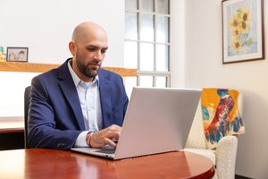 A man in a blue blazer types on a laptop at a wooden table in a room with a window and framed sunflower painting.