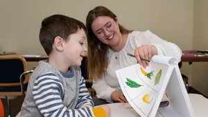 A woman with glasses smiles as she shows a boy a flip chart with colorful vegetable illustrations.