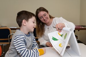 A woman with glasses smiles as she shows a boy a flip chart with colorful vegetable illustrations.