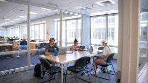 Three students are studying at a large table in a bright academic study room, each focused on their laptop.