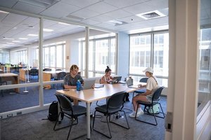Three students are studying at a large table in a bright academic study room, each focused on their laptop.
