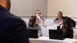 Four students participate in an academic discussion. Two female students smile, and a male student thoughtfully observes