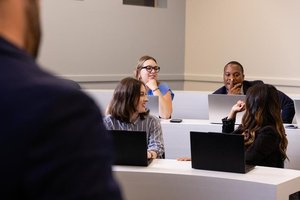 Four students participate in an academic discussion. Two female students smile, and a male student thoughtfully observes