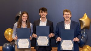Three young adults stand smiling against a dark blue wall, each holding an award certificate with balloons behind them.