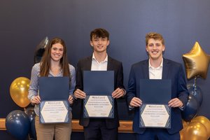 Three young adults stand smiling against a dark blue wall, each holding an award certificate with balloons behind them.