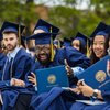 A group of graduates in blue caps and gowns sit outdoors holding diplomas, with one man smiling widely at the camera.