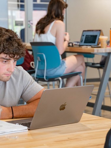 A young man wearing earbuds studies intently on a laptop at a table while two women talk at a table behind him.
