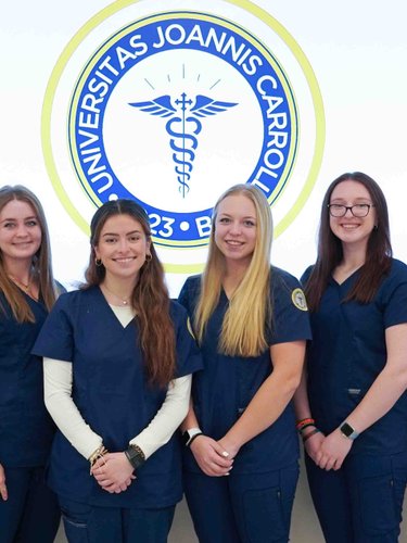 Ten people wearing navy blue medical scrubs stand smiling in a row in front of a large circular university emblem on a white.