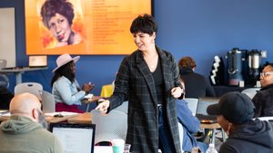 A woman in a checkered coat smiles and points while interacting with seated people in a classroom with laptops and a large.