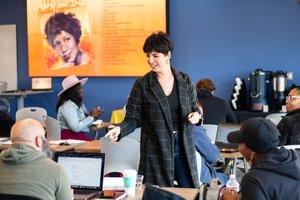 A woman in a checkered coat smiles and points while interacting with seated people in a classroom with laptops and a large.