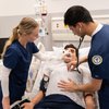 Two medical students in blue scrubs attend to a patient lying in a hospital bed with oxygen mask and blood pressure cuff.