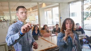 Two students write equations on a glass wall while three others watch from a table behind them.