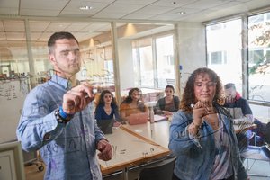 Two students write equations on a glass wall while three others watch from a table behind them.