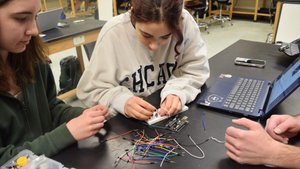 Two students work together connecting wires to a breadboard on a table with a laptop and electronic components.
