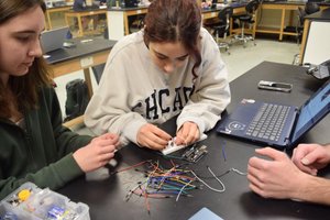 Two students work together connecting wires to a breadboard on a table with a laptop and electronic components.