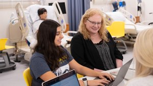 Three people sit at a table with laptops in a room containing hospital beds and medical equipment.