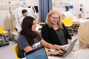 Three people sit at a table with laptops in a room containing hospital beds and medical equipment.