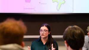 A woman wearing glasses speaks and gestures with her hands in front of a projected colorful map while others listen.