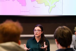 A woman wearing glasses speaks and gestures with her hands in front of a projected colorful map while others listen.