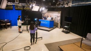 Two people sit behind a news desk while two others operate cameras in a studio with lights and black curtains.
