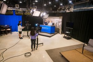 Two people sit behind a news desk while two others operate cameras in a studio with lights and black curtains.