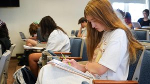 A young woman with long hair writes in a notebook while sitting in a classroom with other students.