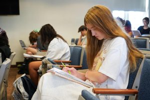 A young woman with long hair writes in a notebook while sitting in a classroom with other students.
