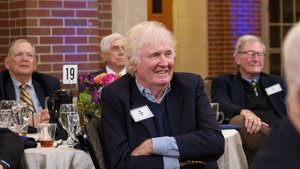 A smiling alumnus with white hair, wearing a navy blazer, sits with crossed arms at a table