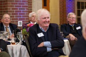 A smiling alumnus with white hair, wearing a navy blazer, sits with crossed arms at a table