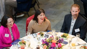 Three alumni share laughter and smiles while enjoying a meal together at a round table with a floral centerpiece during