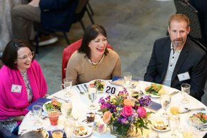 Three alumni share laughter and smiles while enjoying a meal together at a round table with a floral centerpiece during
