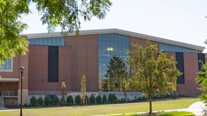 A large brick building with tall glass windows is seen behind a green lawn and a tree under a clear blue sky.
