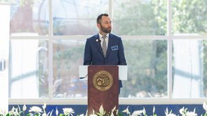 A man speaks at a podium bearing a university seal, with white flowers at its base. He wears a blue suit and tie.