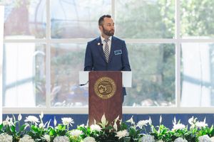 A man speaks at a podium bearing a university seal, with white flowers at its base. He wears a blue suit and tie.