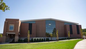 Outside of the athletic center, a brick building with a wall of windows, newly planted saplings stand in the grass.