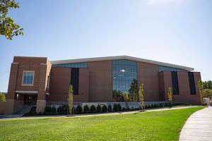 Outside of the athletic center, a brick building with a wall of windows, newly planted saplings stand in the grass.