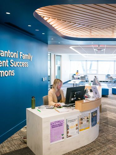 A woman works at a reception desk inside a modern student commons with blue walls and seating areas.