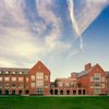 A large red brick building with multiple windows and arched doorways stands under a partly cloudy sky above a green lawn.