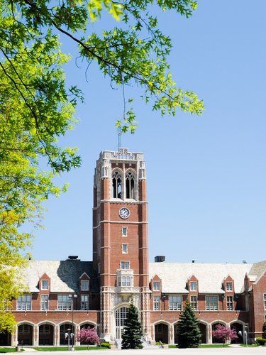Campus view of a brick building with a tower and a clock, set against a clear blue sky and green trees.