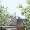 A brick building with many windows and a white cupola is partially framed by blooming white flowers on trees.