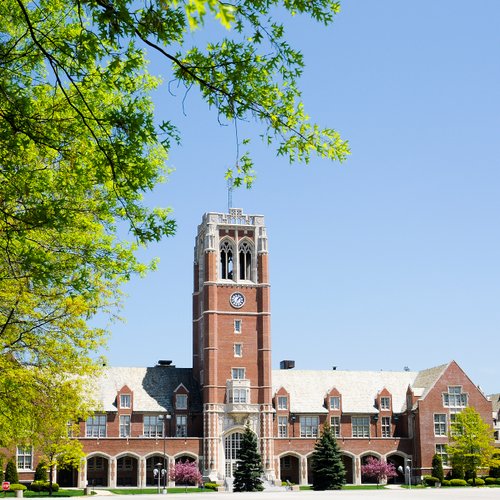 A large brick building with a tall clock tower stands behind green trees under a clear blue sky.