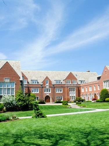 A large red brick building with multiple chimneys and white-framed windows stands behind a green lawn under a blue sky.