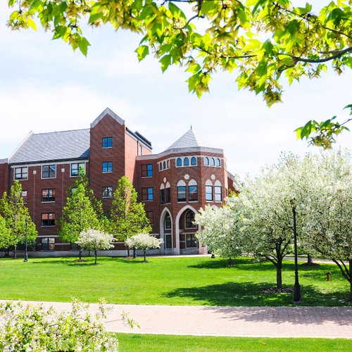 A red brick building with arched windows stands behind a green lawn dotted with blooming white trees under a partly cloudy.