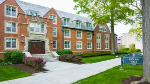 A brick building with multiple windows and a central entrance labeled Dolan Hall stands beside a sidewalk and green lawn.
