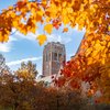 A tall brick clock tower rises behind trees with bright orange and red autumn leaves under a partly cloudy sky.